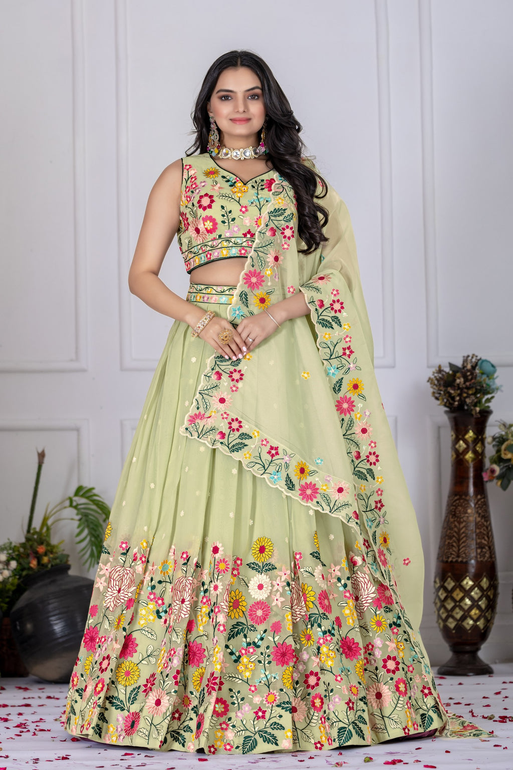 A woman models a light green embroidered Indian ethnic outfit, featuring a sleeveless crop top, a flared skirt with colorful floral patterns, and a matching dupatta, set against a white paneled wall background.